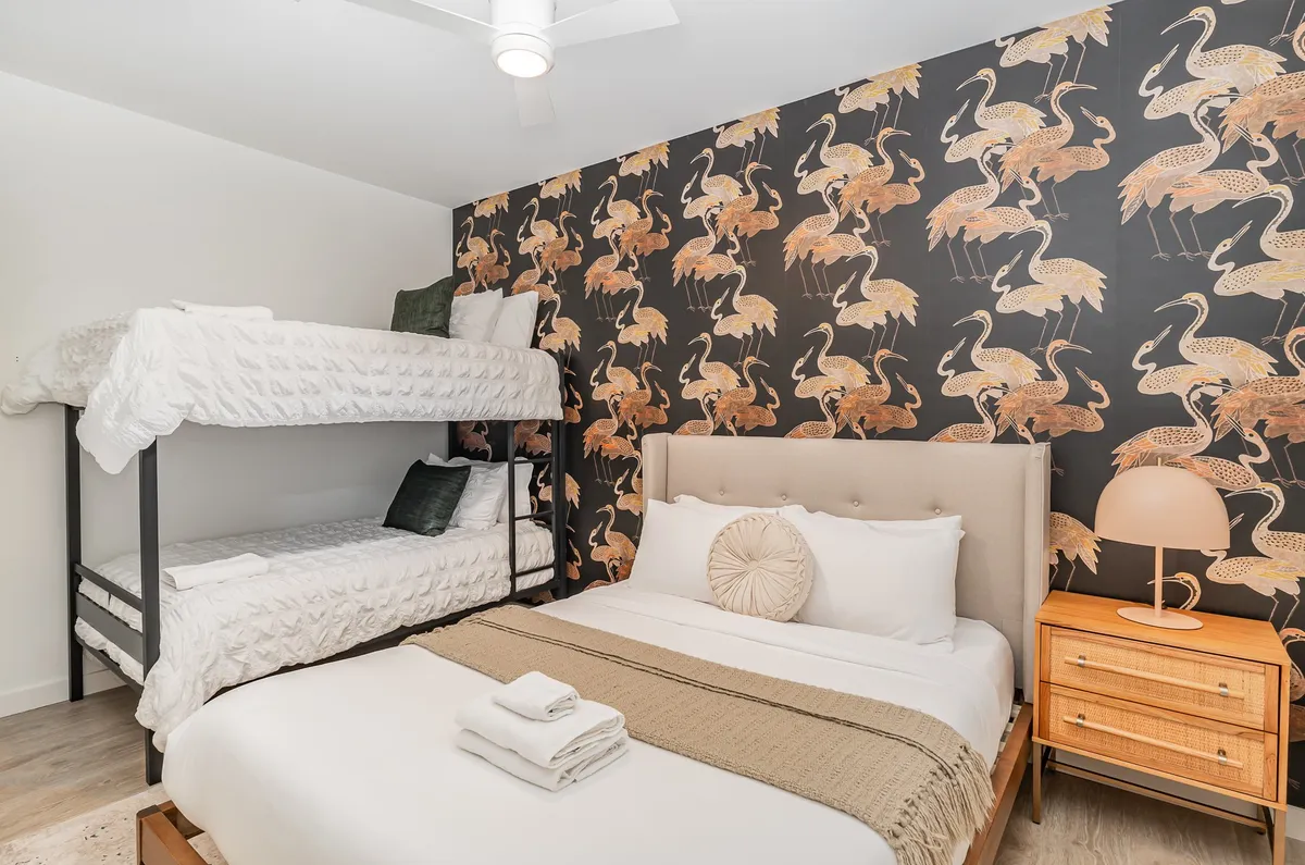 This image displays a bedroom with a queen bed and a bunk bed, playful decorative wallpaper, a nightstand with a lamp, and fun accents that make the room inviting for families or groups. The space feels lively and themed, ideal for late-night giggles and cozy sleepovers.