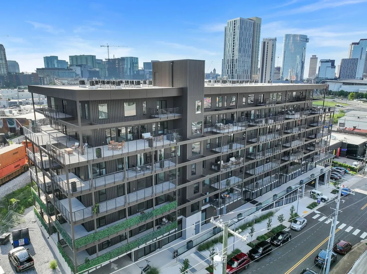 A modern apartment building with lush balconies overlooks a vibrant city skyline. The architecture is sleek and urban, with greenery accenting the building, and the backdrop features Nashville's recognizable downtown. The scene evokes a strong sense of city living and convenience.