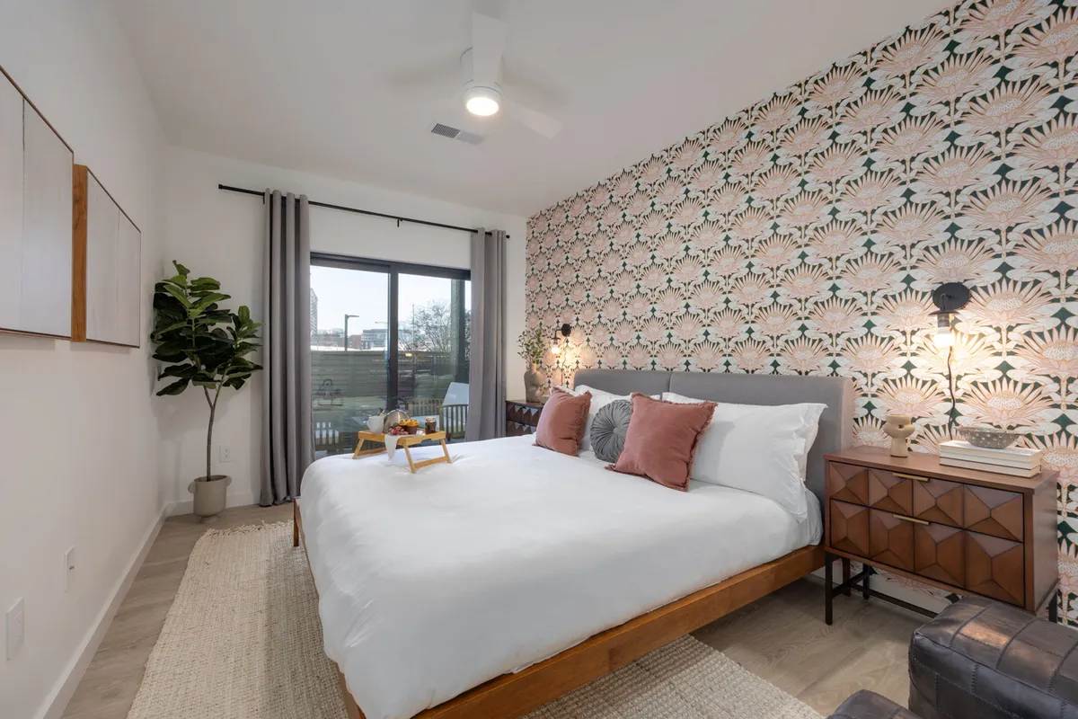 This image showcases a bedroom with a bed adorned with decorative pillows, a bold floral accent wall, a large window flooding the space with natural light, and a side table with a lamp. The playful, nature-inspired theme is visually present, aligning with the whimsical atmosphere described in the section.