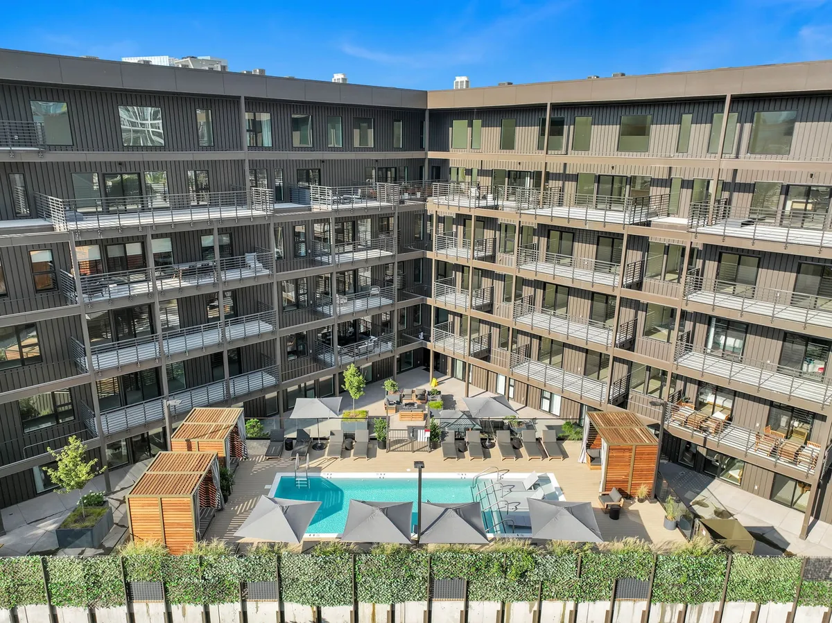 A view of a resort-style swimming pool surrounded by lounge chairs, landscaped greenery, outdoor seating areas, and balconies overlooking the space. The scene exudes a relaxed, social atmosphere with multiple amenities visible.