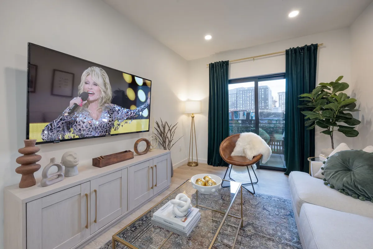 The image showcases a stylish, light-filled living room with a plush sofa facing a large television playing a live musical performance. Green curtains add a pop of color and blend with the indoor plant, tying in the property's jungle-inspired theme. The decorative coffee table offers space for snacks or drinks, making the area ideal for relaxation or socializing.