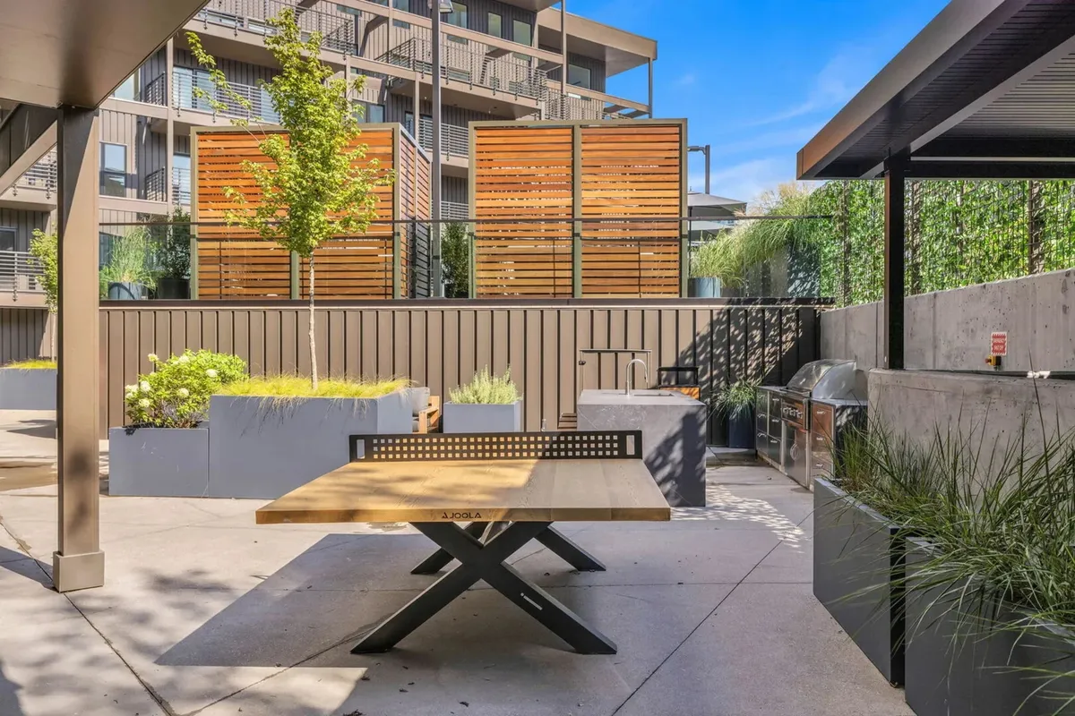 An inviting exterior communal BBQ area with a wooden dining table, grill, lush green plants, and decorative privacy screens. The setup is perfect for outdoor dining, barbecuing, and socializing with friends under the open sky.