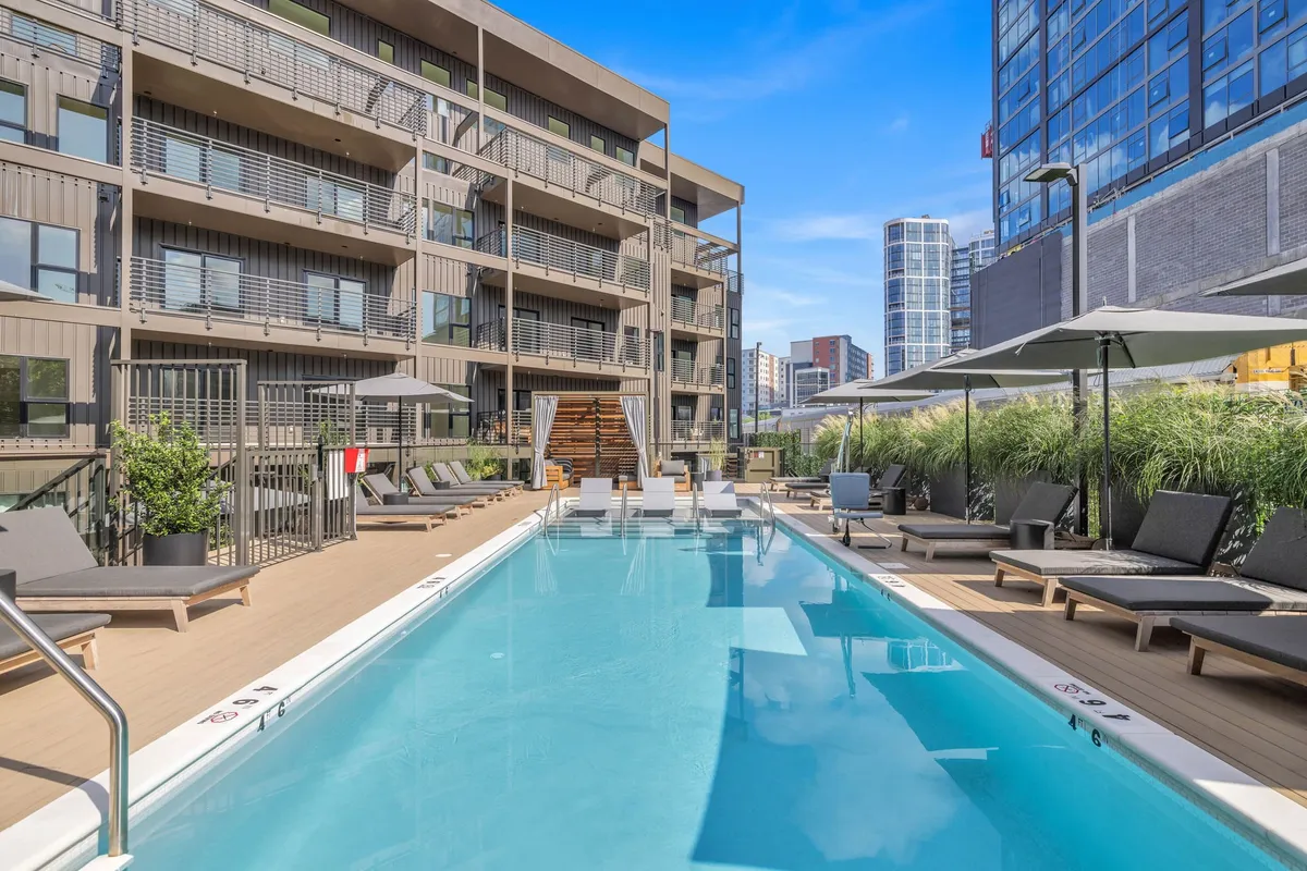 A vibrant exterior shot featuring the swimming pool surrounded by lounge chairs and landscaped greenery, with the building's facade in the background. People are depicted swimming, sunbathing, and socializing beside the water, creating a relaxed yet lively resort-style atmosphere.