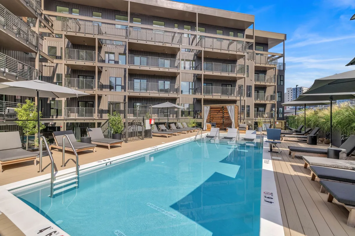 A sparkling outdoor swimming pool surrounded by lounge chairs, balconies, greenery, and umbrellas. The tranquil scene invites relaxation and family fun, suggesting leisure time for all ages within the property itself.