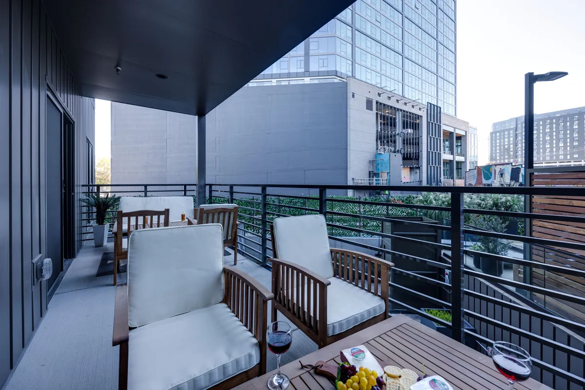 A private balcony setup with two wooden chairs, a small table topped with snacks, a glass of wine, a potted plant, and an urban city backdrop. The scene is inviting and emphasizes relaxation while still being very much part of the city environment.